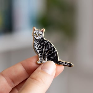 Puede incluir: Un pin de esmalte de un gato atigrado plateado con un contorno dorado. El gato está sentado con la cola enroscada alrededor de sus patas.