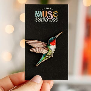 Op de afbeelding: Een kolibrie-speld met een gouden omtrek, rode keel en groene en blauwe accenten wordt getoond op een zwarte kaart. De kaart heeft de tekst "THE GRAY MUSE" in kleurrijke letters. De speld wordt door een hand vastgehouden.