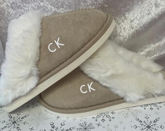 Può includere: Un paio di pantofole in camoscio beige con finiture in ecopelliccia bianca. Le pantofole hanno le lettere "CK" in bianco sul lato. Le suole sono bianche. Su una superficie a motivi di fiocchi di neve.