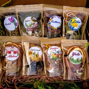 May include: A collection of eight clear pouches filled with dried herbs and botanicals, each sealed with a round sticker. The stickers feature colorful illustrations and the names "Imbolc", "Ostara", "Beltane", "Litha", "Mabon", "Lughnasadh", "Samhain", and "Yule". The pouches are arranged in a brown box.