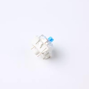 May include: A single mechanical keyboard switch with a white base, clear housing, and a blue stem. The switch has two gold-colored pins extending from the bottom. The clear housing allows a view of the internal components.