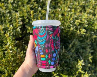 May include: A white to-go cup with a black, colorful, geometric patterned fabric sleeve. The sleeve has a variety of colors including red, blue, green, yellow, and pink. A straw is sticking out of the top of the cup.