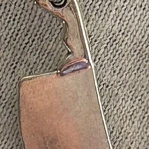 May include: A silver charm in the shape of a chef's knife. The knife has a rounded handle and a sharp blade.