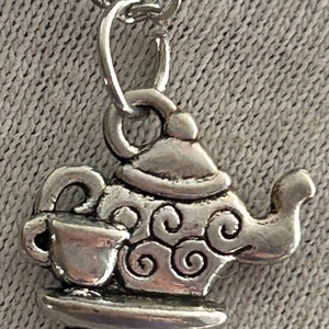May include: Silver charm necklace featuring a teapot and cup design. The teapot has a swirl pattern and a lid. The cup is on a small saucer.