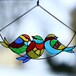 May include: Three stained glass birds hanging from a chain. The birds are blue, green, yellow, red, and orange. The birds are in a curved formation.