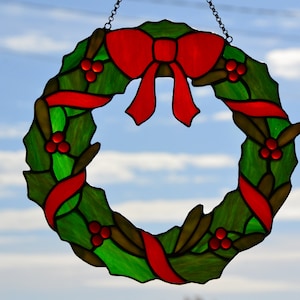May include: A stained glass Christmas wreath with a red bow and green holly leaves. The wreath is hanging from a chain.