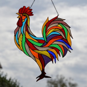May include: A colorful stained glass rooster with a red comb and wattle, hanging from a chain. The rooster's tail feathers are a rainbow of colors, including red, orange, yellow, green, blue, and purple.