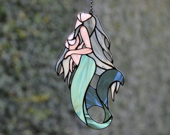 Op de afbeelding: Een hangende zeemeermin-versiering van gekleurd glas met een turquoise staart, grijs haar en een roze bovenlichaam.