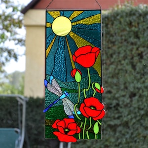 May include: Stained glass panel featuring a sunny sky, red poppies, and a dragonfly. The sun is yellow, with golden rays. The poppies are bright red, and the dragonfly has blue and silver wings. The panel is rectangular and hangs from a chain.