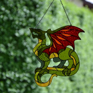 May include: A stained glass suncatcher shaped like a dragon with a green body and textured red and orange wings. The dragon is constructed from various pieces of colored glass held together by dark metal solder lines and hangs from a thin metal chain.
