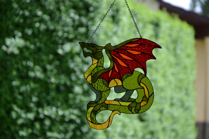 May include: A stained glass suncatcher shaped like a dragon with a green body and textured red and orange wings. The dragon is constructed from various pieces of colored glass held together by dark metal solder lines and hangs from a thin metal chain.