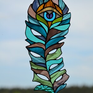 May include: Stained glass feather with an eye design. The feather features shades of blue, green, brown, and orange. The eye has a blue iris and a purple pupil. The piece is suspended by a black chain.