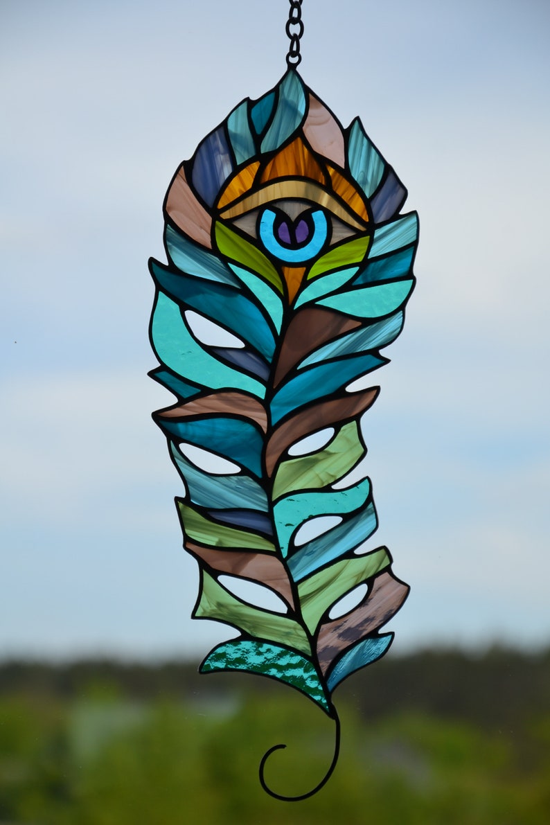 May include: Stained glass feather with an eye design. The feather features shades of blue, green, brown, and orange. The eye has a blue iris and a purple pupil. The piece is suspended by a black chain.