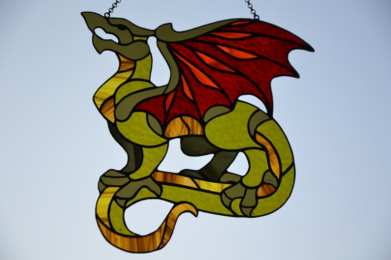 May include: A stained glass suncatcher shaped like a dragon with a green body, yellow accents, and large red and orange wings. The dragon is depicted in a standing pose with its tail curled, featuring dark lead lines separating the glass pieces.