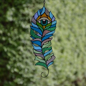 May include: Stained glass feather with an eye design. The feather features shades of blue, green, and purple, with an eye at the top. The artwork is suspended by a chain, showcasing a unique decorative piece.