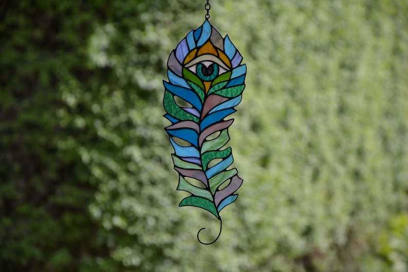 May include: Stained glass feather with an eye design. The feather features shades of blue, green, and purple, with an eye at the top. The artwork is suspended by a chain, showcasing a unique decorative piece.