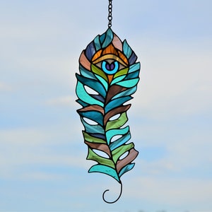May include: A stained glass feather-shaped suncatcher with a chain for hanging. The design features an eye at the top and is composed of teal, brown, green, and blue glass pieces. The background is a clear, light blue sky.