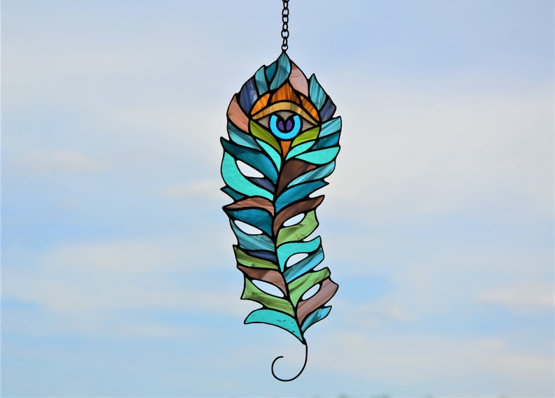 May include: A stained glass feather-shaped suncatcher with a chain for hanging. The design features an eye at the top and is composed of teal, brown, green, and blue glass pieces. The background is a clear, light blue sky.
