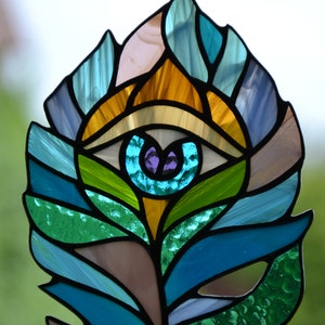 May include: Stained glass feather with an eye design. The feather features a variety of colors, including blue, green, yellow, and purple. The eye is in the center of the feather, with a purple iris and a white and black outline.
