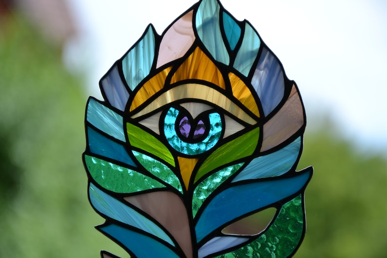 May include: Stained glass feather with an eye design. The feather features a variety of colors, including blue, green, yellow, and purple. The eye is in the center of the feather, with a purple iris and a white and black outline.