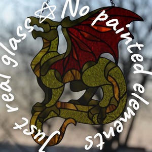 May include: A stained glass suncatcher shaped like a dragon with textured olive green body segments and deep red wings. The piece features dark leaded lines and includes the text "Just real glass ☆ No painted elements" and a logo for Glass IC Studio.