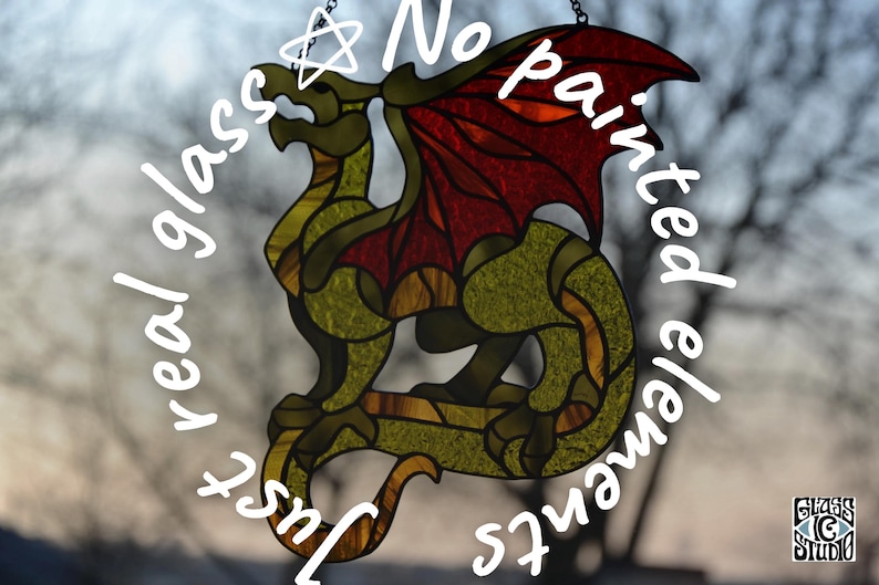 May include: A stained glass suncatcher shaped like a dragon with textured olive green body segments and deep red wings. The piece features dark leaded lines and includes the text "Just real glass ☆ No painted elements" and a logo for Glass IC Studio.