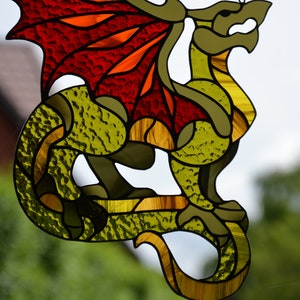 May include: A stained glass suncatcher shaped like a dragon with textured olive green glass for the body and vibrant red and orange glass for the wings. The dragon is suspended by a thin metal chain and features dark leaded lines outlining its form.