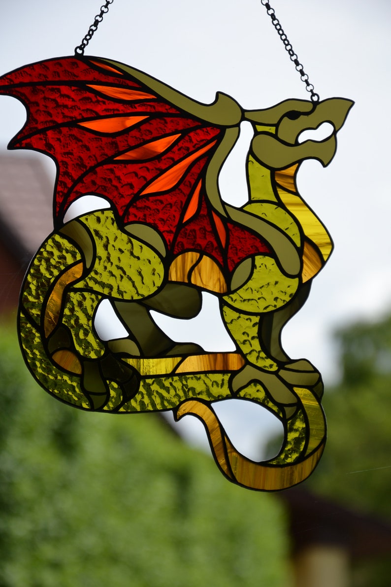 May include: A stained glass suncatcher shaped like a dragon with textured olive green glass for the body and vibrant red and orange glass for the wings. The dragon is suspended by a thin metal chain and features dark leaded lines outlining its form.
