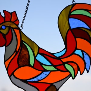 May include: A stained glass rooster with a red, orange, yellow, green, blue, and gray color scheme. The rooster is hanging from a chain.