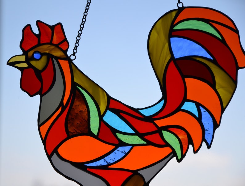 May include: A stained glass rooster with a red, orange, yellow, green, blue, and gray color scheme. The rooster is hanging from a chain.