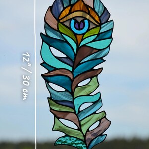 May include: Stained glass feather, 12 inches (30 cm) long and 4.5 inches (11 cm) wide, with an eye design. The feather features shades of blue, green, brown, and orange. The Glass G Studio logo is in the bottom right corner.