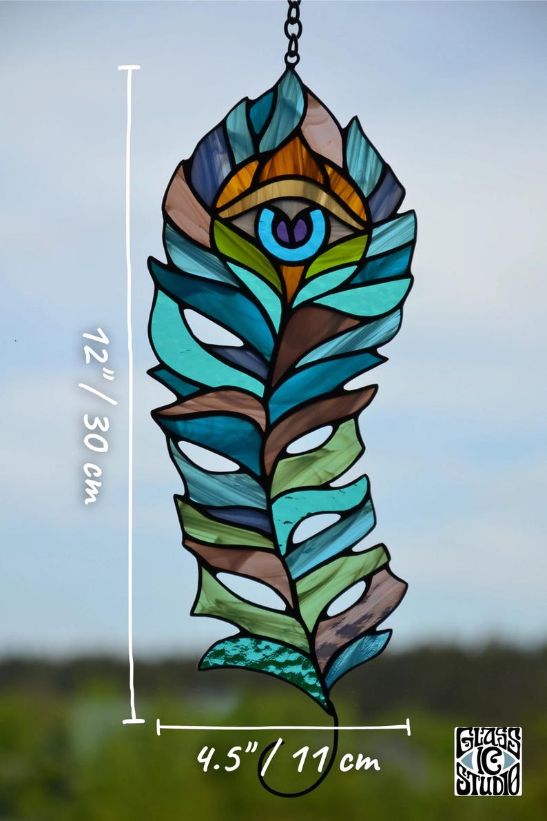 May include: Stained glass feather, 12 inches (30 cm) long and 4.5 inches (11 cm) wide, with an eye design. The feather features shades of blue, green, brown, and orange. The Glass G Studio logo is in the bottom right corner.