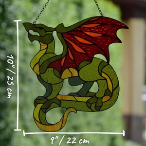 May include: A stained glass dragon suncatcher featuring green, yellow, and red glass segments. The dragon measures 10 inches in height and 9 inches in width. It is suspended by a metal chain and displays a Glass IC Studio logo in the corner.