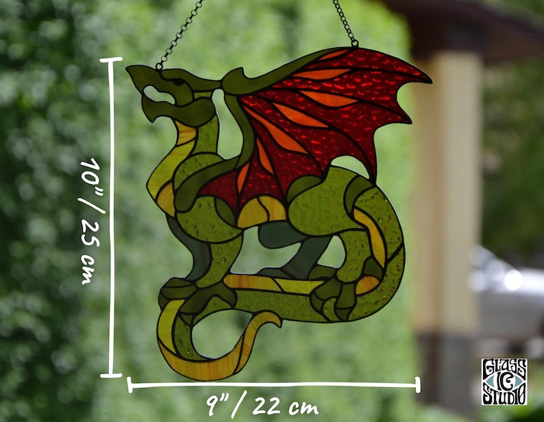 May include: A stained glass dragon suncatcher featuring green, yellow, and red glass segments. The dragon measures 10 inches in height and 9 inches in width. It is suspended by a metal chain and displays a Glass IC Studio logo in the corner.