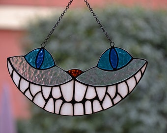 May include: A stained glass Cheshire Cat window hanging with a wide, toothy grin, blue eyes, and an orange nose.