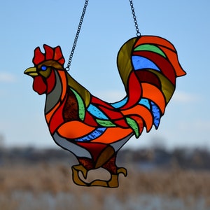 May include: A stained glass rooster with a red, orange, yellow, green, blue, and brown color scheme. The rooster is hanging from a chain.