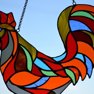 May include: A stained glass rooster with a red, orange, yellow, green, and blue color scheme. The rooster is hanging from a chain.