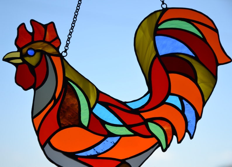May include: A stained glass rooster with a red, orange, yellow, green, and blue color scheme. The rooster is hanging from a chain.