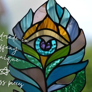May include: A stained glass feather in shades of blue, green, gold, and purple, created using the Tiffany technique. The artwork features an eye design at the center and the text "Handmade in Tiffany Technique" and "50 glass pieces".