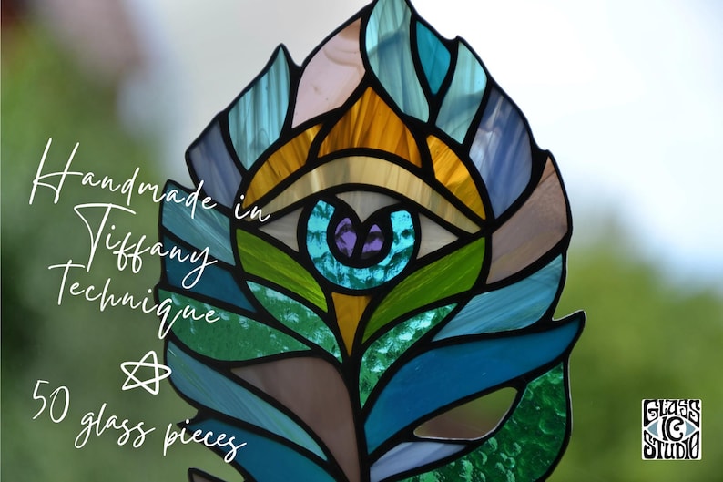 May include: A stained glass feather in shades of blue, green, gold, and purple, created using the Tiffany technique. The artwork features an eye design at the center and the text "Handmade in Tiffany Technique" and "50 glass pieces".