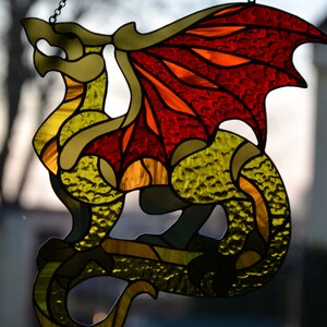 May include: A stained glass suncatcher shaped like a dragon with a textured yellow and green body and large, vibrant red and orange wings. The piece is constructed with dark metal lead lines and includes a small metal chain for hanging purposes.