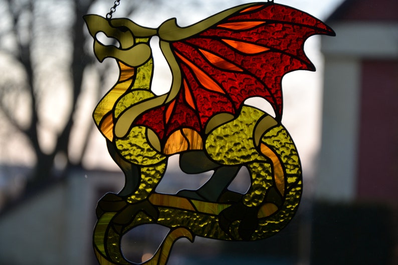 May include: A stained glass suncatcher shaped like a dragon with a textured yellow and green body and large, vibrant red and orange wings. The piece is constructed with dark metal lead lines and includes a small metal chain for hanging purposes.
