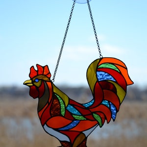 May include: A stained glass rooster with a red, orange, yellow, green, and blue color scheme. The rooster is hanging from a chain and a suction cup.