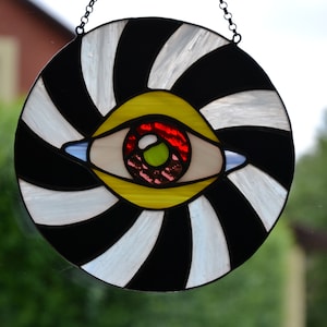 May include: A stained glass window hanging with a black and white spiral design and a large eye in the center. The eye is yellow, red, and green.