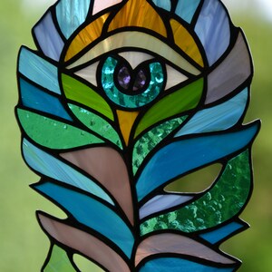 May include: Stained glass feather with an eye design. The feather features a vibrant array of colors, including shades of blue, green, yellow, and purple. The eye is in the center of the feather, with a teal iris and a purple pupil. The feather is outlined in black.