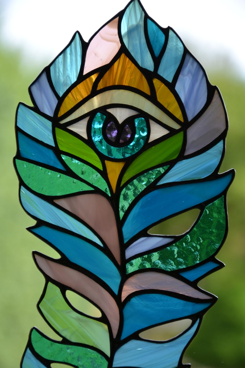 May include: Stained glass feather with an eye design. The feather features a vibrant array of colors, including shades of blue, green, yellow, and purple. The eye is in the center of the feather, with a teal iris and a purple pupil. The feather is outlined in black.