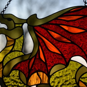 May include: A stained glass suncatcher shaped like a dragon, featuring a body made of textured yellow and green glass pieces, and large wings constructed from vibrant red and orange glass segments, all held together by dark metal solder lines.