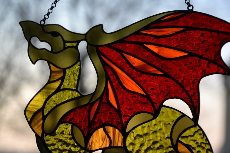 May include: A stained glass suncatcher shaped like a dragon, featuring a body made of textured yellow and green glass pieces, and large wings constructed from vibrant red and orange glass segments, all held together by dark metal solder lines.