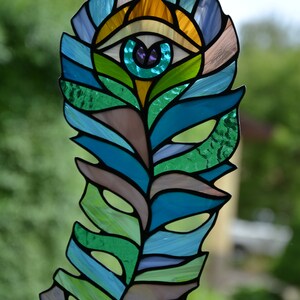 May include: Stained glass feather with an eye design. The feather features shades of blue, green, and brown, with an eye in the center. The eye has a teal iris and a purple pupil. The feather is hanging from a black chain.
