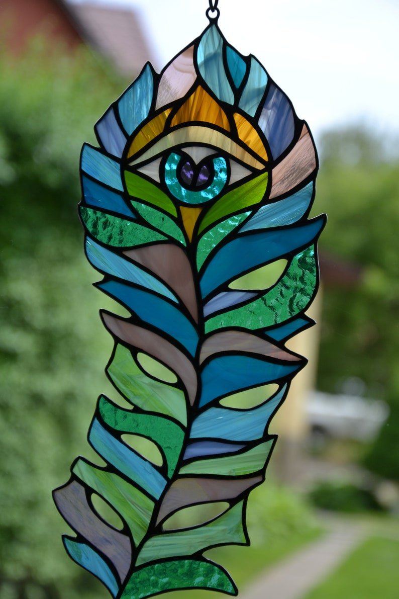 May include: Stained glass feather with an eye design. The feather features shades of blue, green, and brown, with an eye in the center. The eye has a teal iris and a purple pupil. The feather is hanging from a black chain.